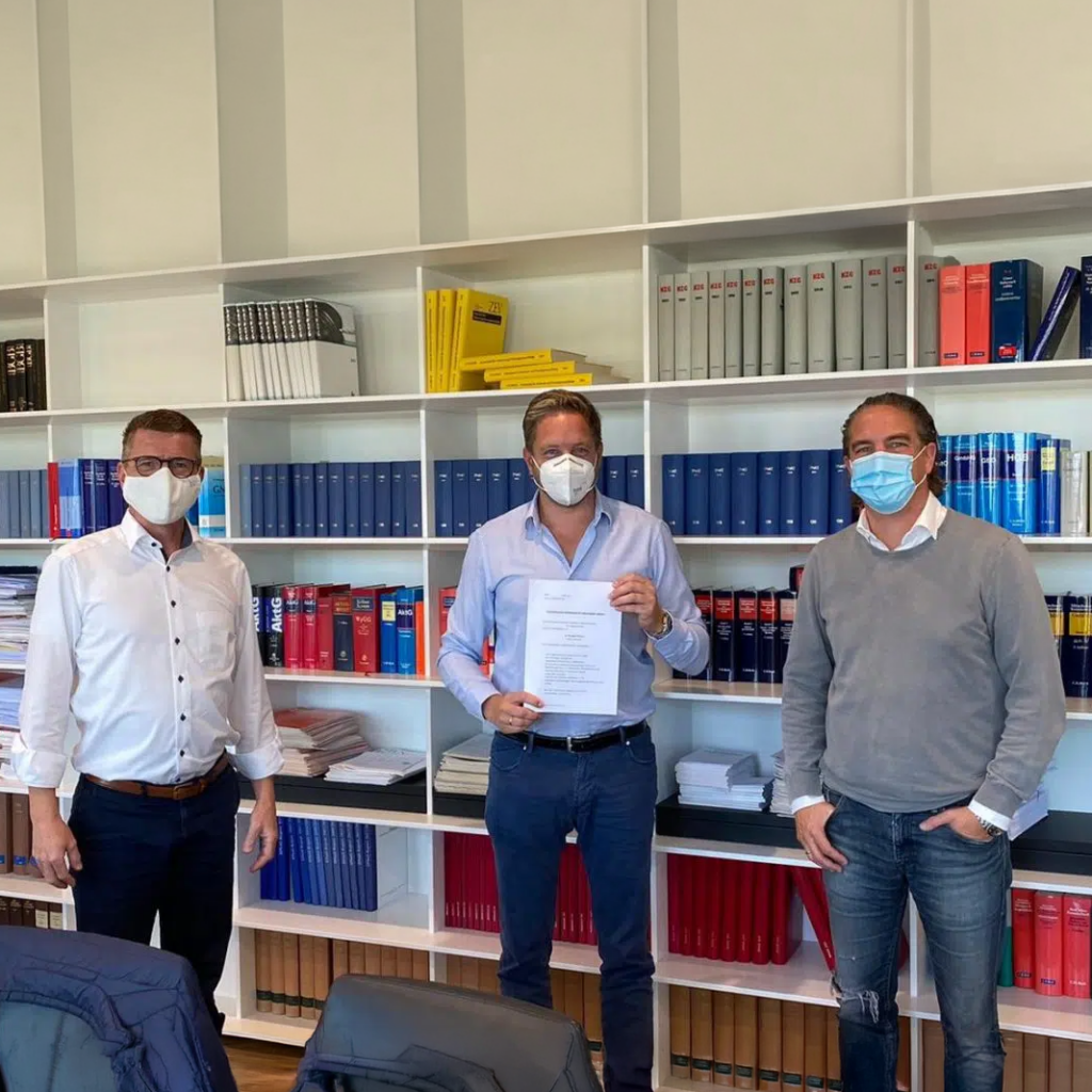 Zu sehen sind drei Männer die vor einem Bücherregal stehen. Der in der Mitte hält die Beteiligungsurkunde in der Hand.
