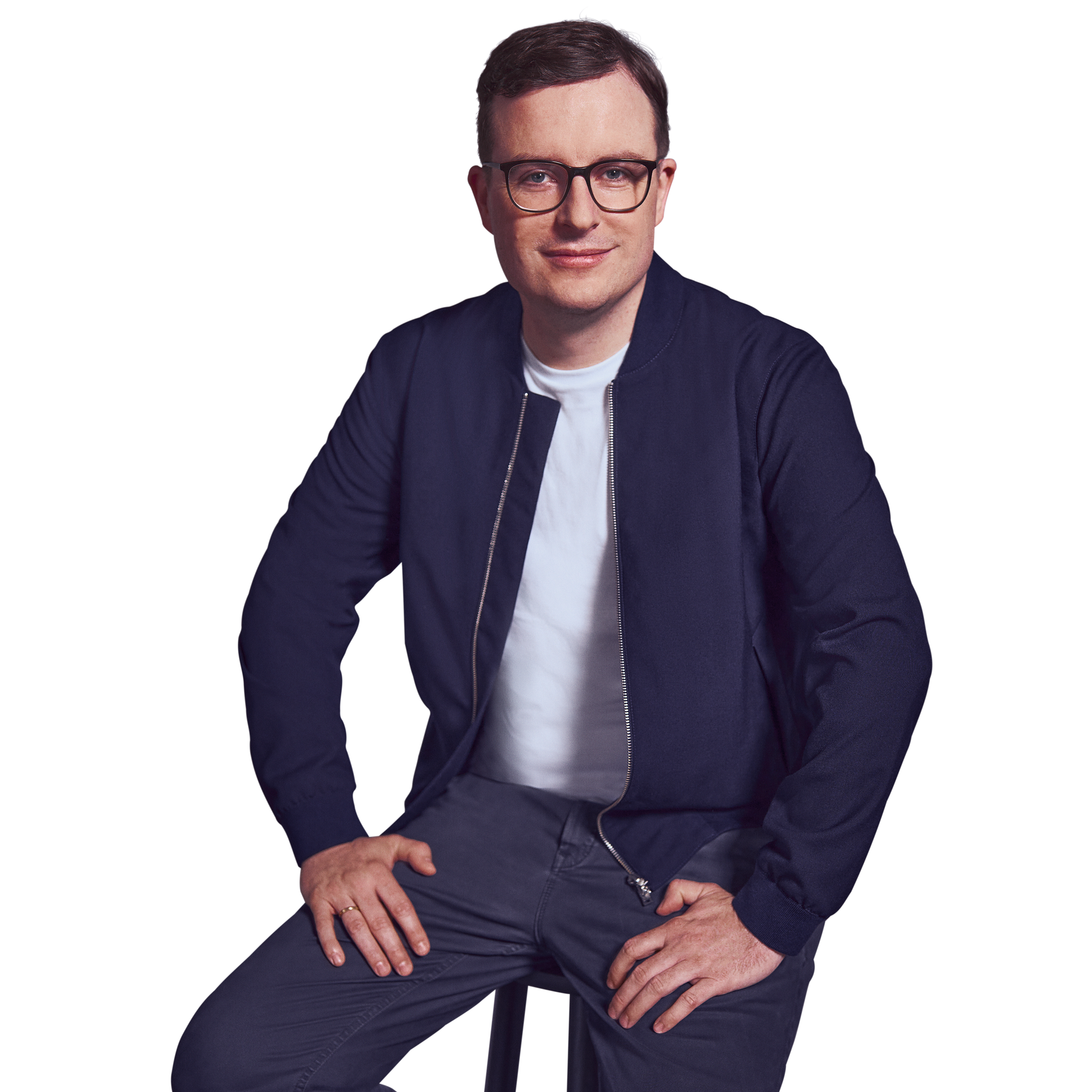 Männliche Person, braune kurze Haare, dunkelblaue Augen mit Brille, lächelnd, trägt ein weißes T-Shirt, eine dunkelblaue Jacke und Hose, sitzend mit beiden Händen auf dem Schoß abstützend