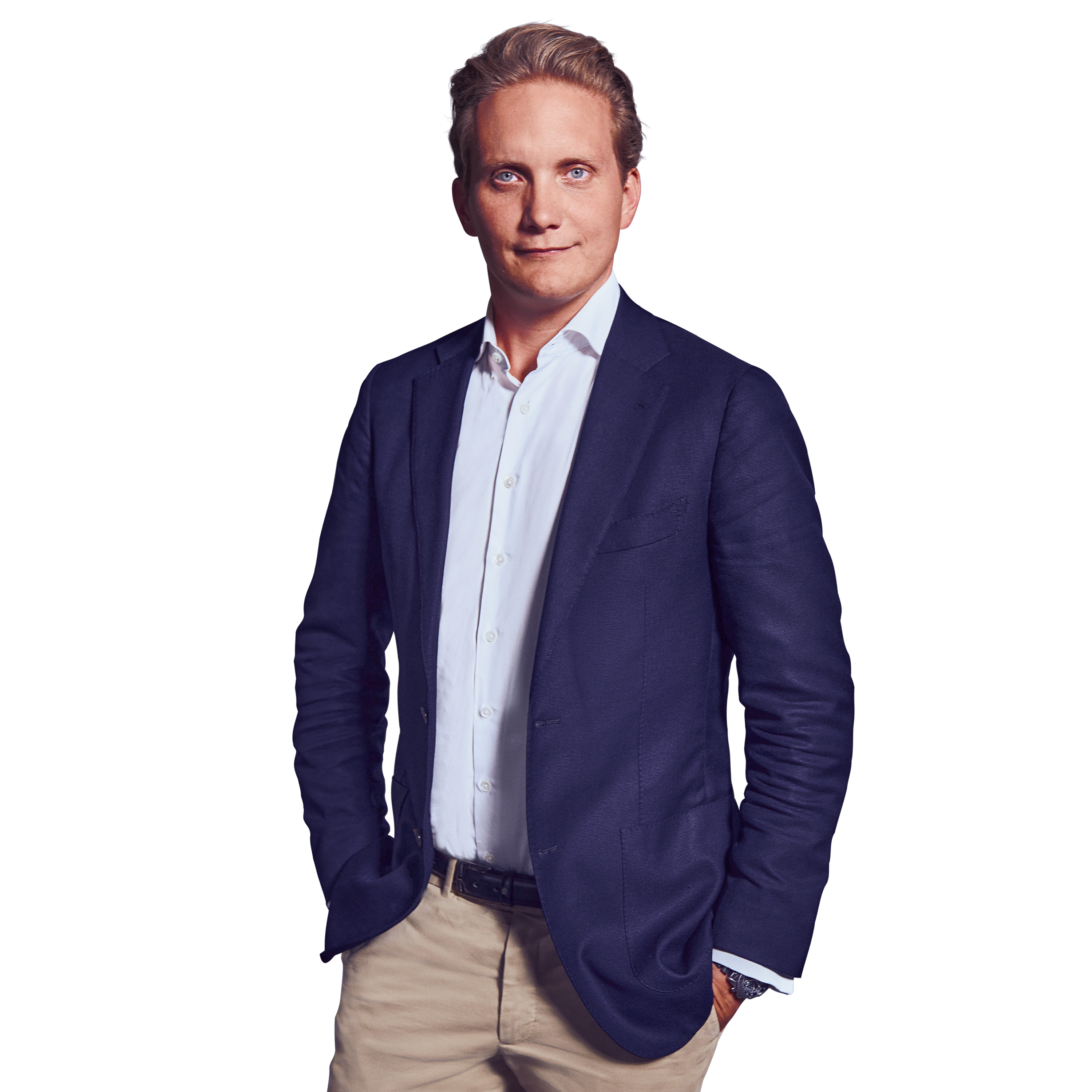 Männliche Person, blonde kurze Haare, blaue Augen, neutraler Gesichtsausdruck, trägt ein weißes Hemd, eine dunkelblaue Jacke und beige Hose, stehend mit beiden Händen in den Hosentaschen