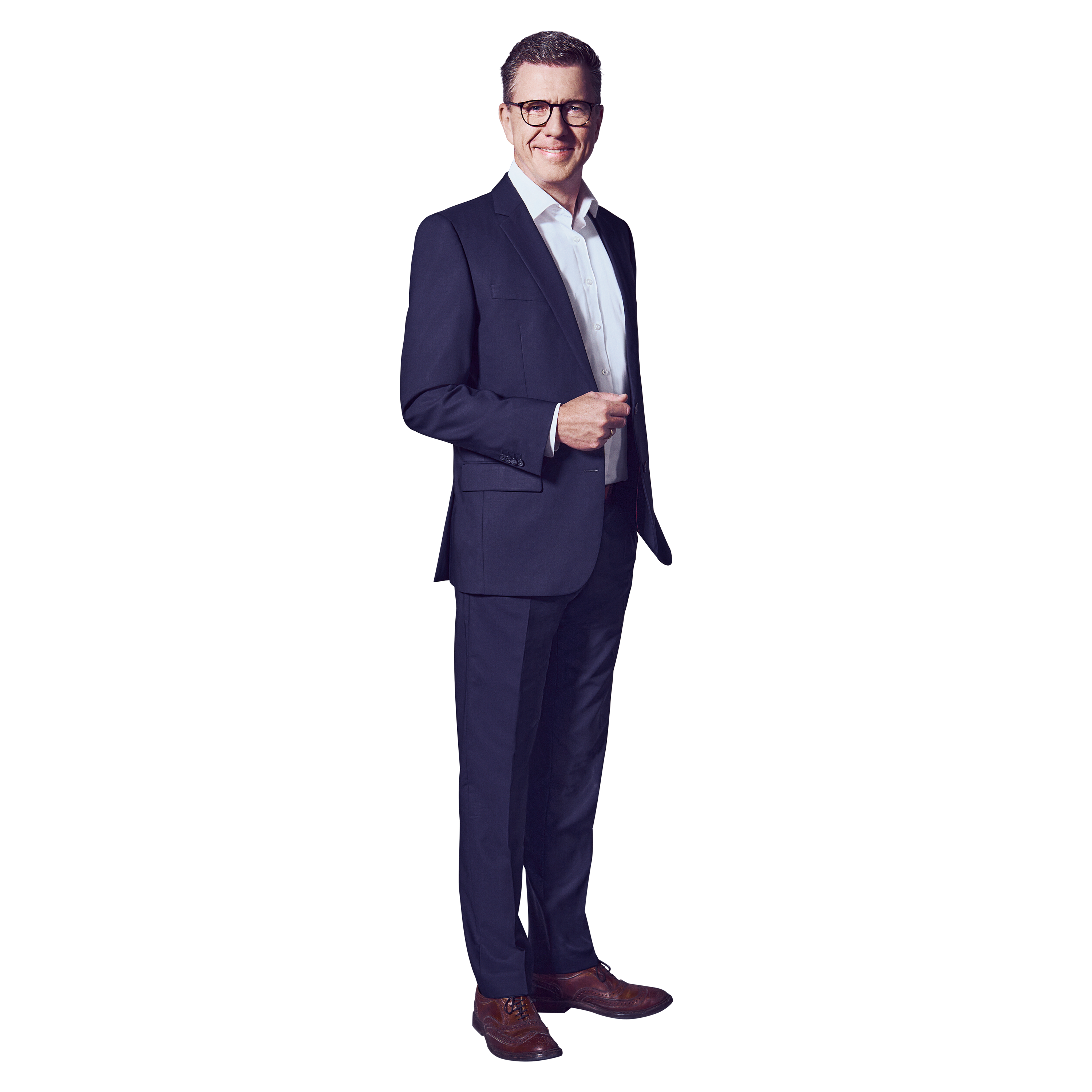 Male, dark blond hair, blue eyes, wearing glasses, smiling, dressed in a white shirt and dark blue suit, brown shoes, standing with his left hand in his pants pocket and his right hand bent at the elbow in front of his upper body