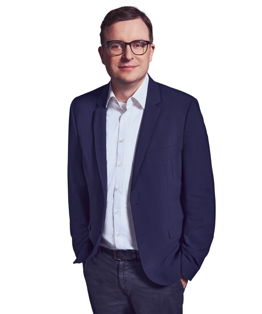Porträtfoto von Bene Budde, Partner bei Kemény Boehme Consultants (KBC). Mann mit kurzen, dunkelbraunen Haaren und einer markanten schwarzen Rechteckbrille blickt freundlich lächelnd in die Kamera. Trägt ein dunkelblaues Sakko über einem weißen Hemd ohne Krawatte sowie eine dunkle Jeanshose und einen schwarzen Ledergürtel, beide Hände locker in den Hosentaschen, entspannte und zugängliche Haltung.