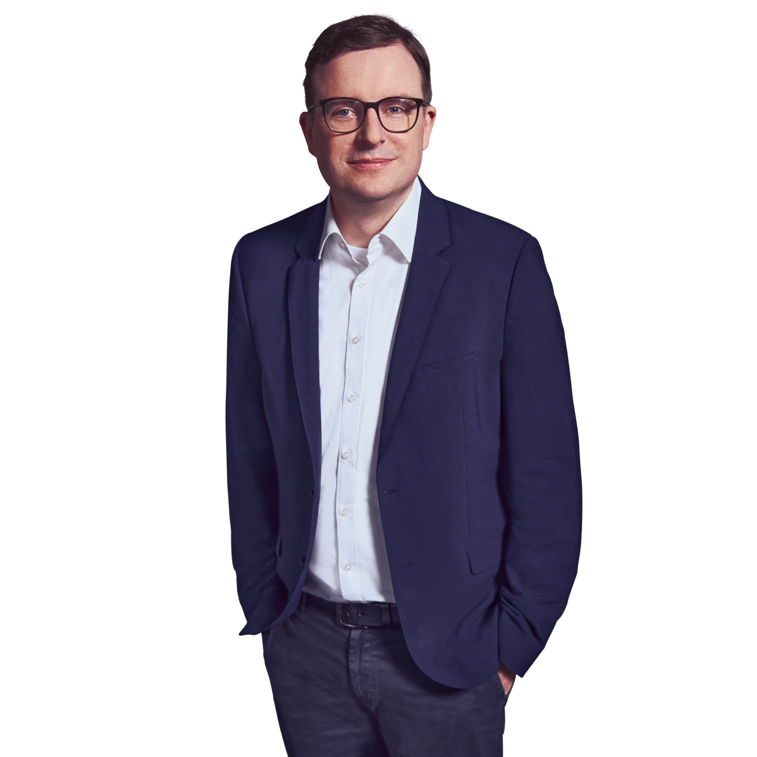 Porträtfoto von Bene Budde, Partner bei Kemény Boehme Consultants (KBC). Mann mit kurzen, dunkelbraunen Haaren und einer markanten schwarzen Rechteckbrille blickt freundlich lächelnd in die Kamera. Trägt ein dunkelblaues Sakko über einem weißen Hemd ohne Krawatte sowie eine dunkle Jeanshose und einen schwarzen Ledergürtel, beide Hände locker in den Hosentaschen, entspannte und zugängliche Haltung.
