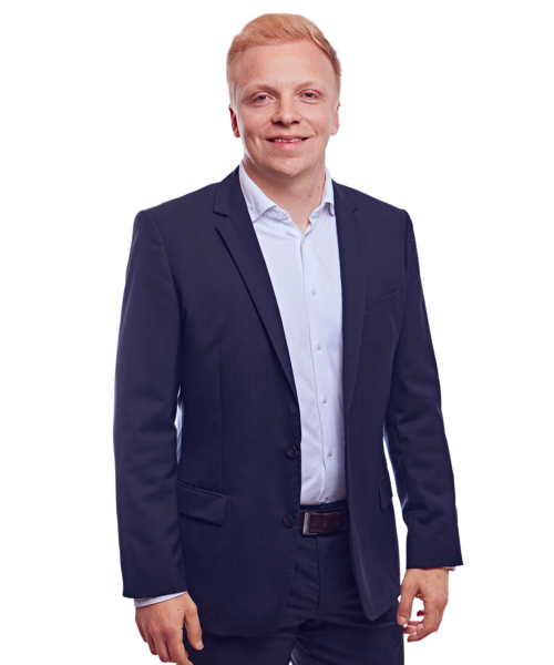 Porträtfoto von Constantin von Unruh, Manager bei Kemény Boehme Consultants (KBC). Mann mit kurzem, hellblondem bis rötlichem Haar blickt freundlich lächelnd in die Kamera. Trägt einen dunklen Blazer über einem hellblauen, offen getragenen Hemd ohne Krawatte sowie einen dunklen Gürtel, Arme locker seitlich am Körper, aufrechte und natürliche Haltung.