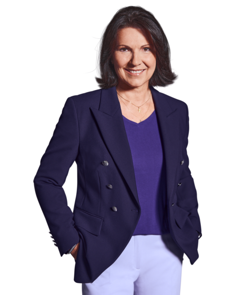 Porträtfoto von Constanze Schöffmann, Director of HR bei Kemény Boehme Consultants (KBC). Frau mit schulterlangem, dunkelbraunem Haar mit grauen Strähnen, blickt freundlich lächelnd in die Kamera. Trägt ein dunkelblaues Doppelreiher-Sakko über einem lilafarbenen V-Ausschnitt-Shirt und eine weiße Hose sowie eine feine goldene Halskette. Hände in den Hosentaschen, souveräne und einladende Haltung.