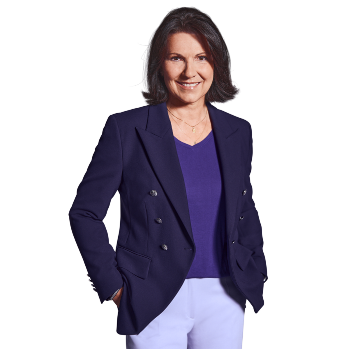 Portrait photo of Constanze Schöffmann, Director of HR at Kemény Boehme Consultants (KBC). A woman with shoulder-length, dark brown hair streaked with gray, looks into the camera with a friendly smile. She is wearing a dark blue double-breasted blazer over a purple V-neck shirt and white pants, as well as a delicate gold necklace. Her hands are in her pockets, and she has a confident and welcoming demeanor.