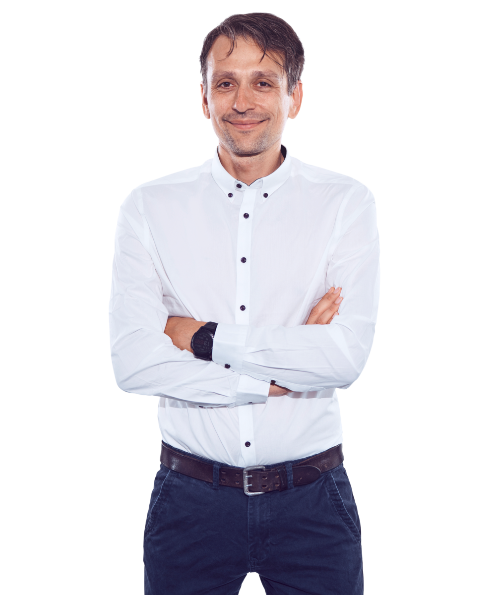 Porträtfoto von Denis Zyulyamov, Head of IT bei Kemény Boehme Consultants (KBC). Mann mit kurzen, dunkelbraunen Haaren und leichtem Bartschatten, blickt verschmitzt lächelnd in die Kamera. Trägt ein weißes Button-down-Hemd mit dunklen Knöpfen und eine dunkelblaue Chinohose mit braunem Ledergürtel sowie eine schwarze Uhr am Handgelenk. Arme verschränkt, entspannte und sympathische Haltung.