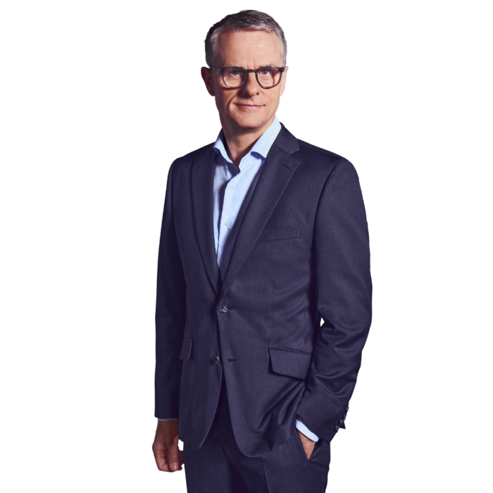 Portrait photo of John Dowling, partner at Kemény Boehme Consultants (KBC). A man with short, graying hair and distinctive dark brown rectangular glasses looks calmly and intently into the camera. He is wearing a dark blue suit with a matching jacket and pants over a light blue shirt without a tie, one hand casually in his pants pocket, the other loosely at his side, with an upright and confident posture.