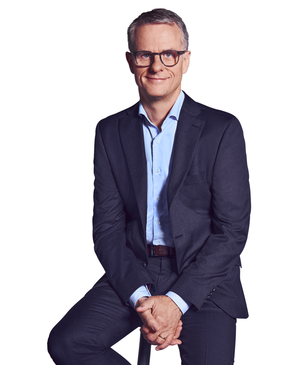Porträtfoto von John Dowling, Partner bei Kemény Boehme Consultants (KBC). Mann mit kurzen, grau-melierten Haaren und markanter dunkelbrauner Rechteckbrille blickt ruhig und konzentriert in die Kamera. Trägt einen dunkelblauen Anzug mit passendem Sakko und Hose über einem hellblauen Hemd ohne Krawatte, eine Hand lässig in der Hosentasche, die andere locker seitlich am Körper, aufrechte und souveräne Haltung.