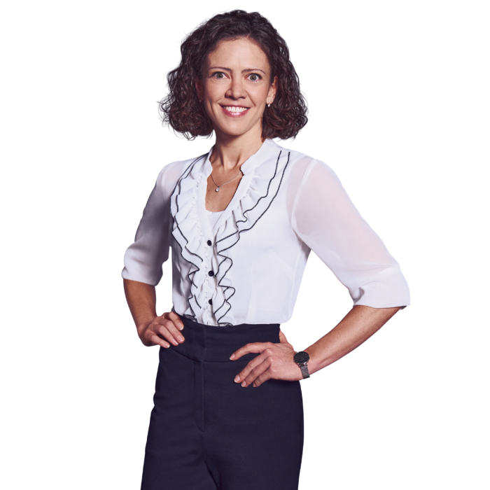 Portrait photo of Larissa Fahrmeier, Director at Kemény Boehme Consultants (KBC). A woman with shoulder-length, dark brown curly hair smiles broadly at the camera. She is wearing an elegant white chiffon blouse with black ruffles and a button placket over dark blue dress pants, as well as a delicate silver necklace and a silver wristwatch; both hands are confidently placed on her hips, and she has an energetic and open posture.