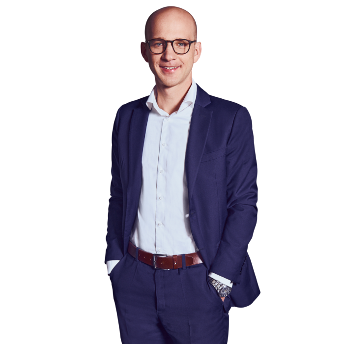 Portrait photo of Martin Betz, Director at Kemény Boehme Consultants (KBC). A man with a shaved head and distinctive dark rectangular glasses, smiling warmly at the camera. He is wearing a dark blue suit with a matching jacket and pants over a light blue shirt without a tie, a cognac-colored leather belt, and a silver wristwatch on his right wrist; both hands are in his pants pockets, and he has an upright and confident posture.