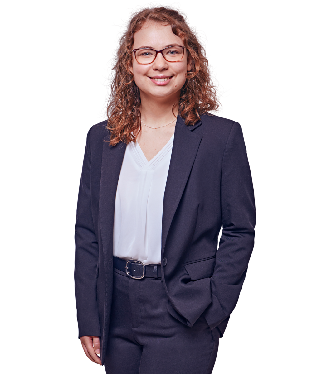 Porträtfoto von Sophie-Theres Altmann, Consultant bei Kemény Boehme Consultants (KBC). Frau mit schulterlagen, lockigen rotbraunen Haaren und einer markanten eckigen Brille mit rotem Rahmen, blickt freundlich lächelnd in die Kamera. Trägt ein dunkelblaues Sakko über einer weißen V-Ausschnitt-Bluse und eine dunkle Hose mit schwarzem Gürtel sowie eine feine Halskette. Eine Hand in der Hosentasche, entspannte und professionelle Haltung.