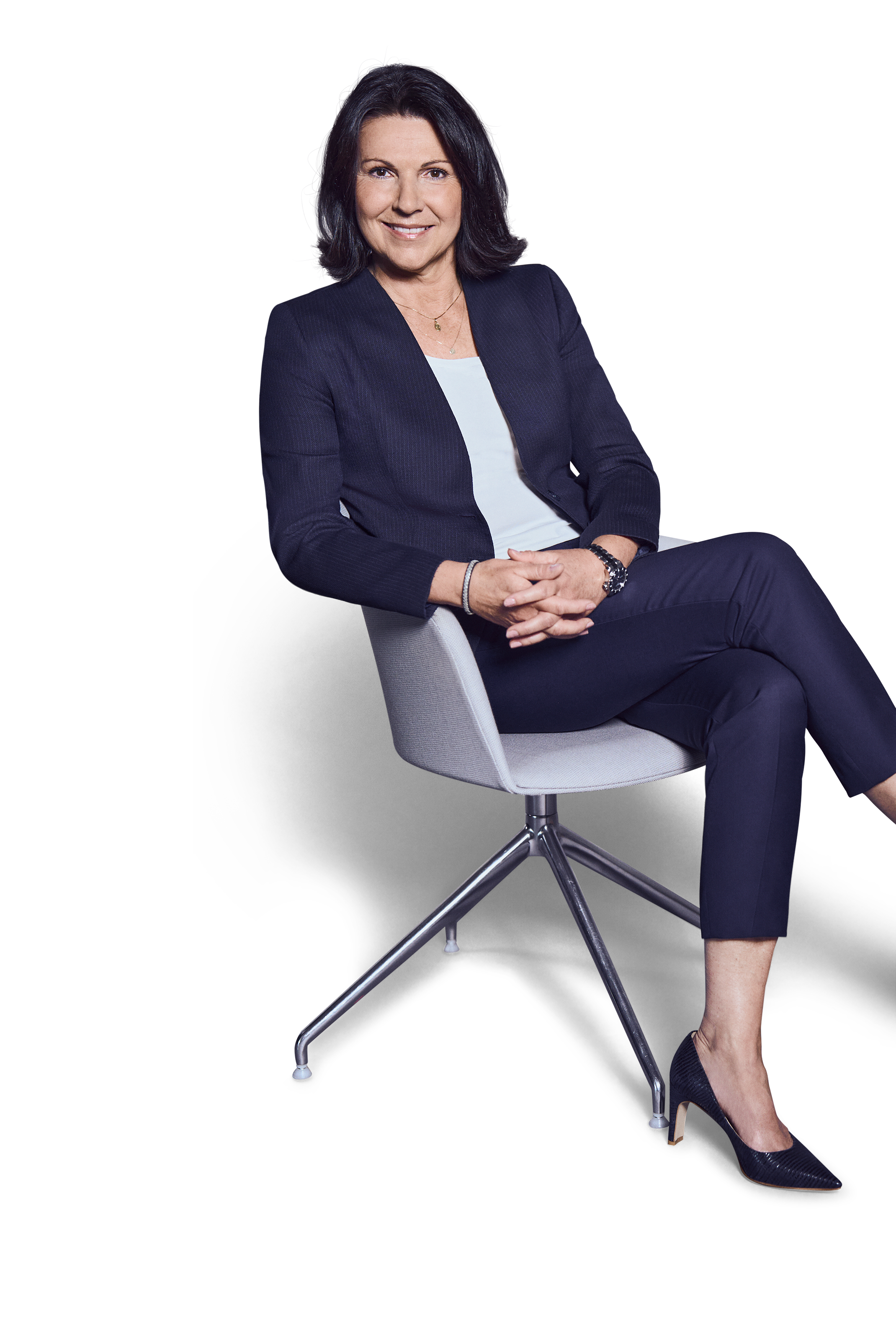 Weibliche Person, schwarze mittellange Haare, braune Augen, lächelnd, trägt ein weißes Oberteil, dunkelblaue Hose und Blazer, sitzend mit überschlagenen Beinen und verschränkten Händen