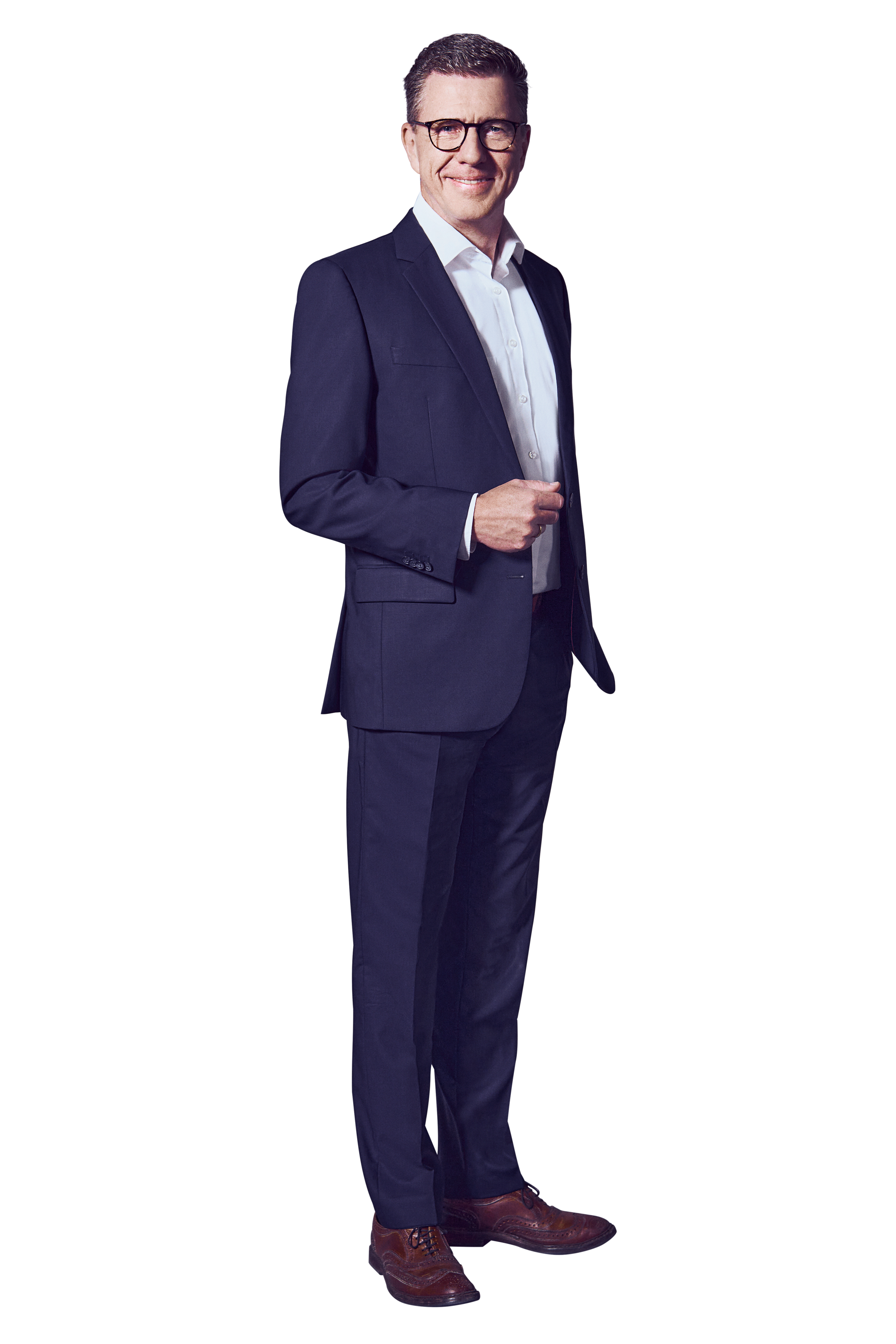 Male, dark blond hair, blue eyes, wearing glasses, smiling, dressed in a white shirt and dark blue suit, brown shoes, standing with his left hand in his pants pocket and his right hand bent at the elbow in front of his upper body