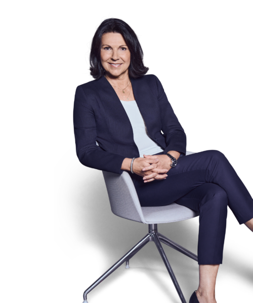 Weibliche Person, schwarze mittellange Haare, braune Augen, lächelnd, trägt ein weißes Oberteil, dunkelblaue Hose und Blazer, sitzend mit überschlagenen Beinen und verschränkten Händen