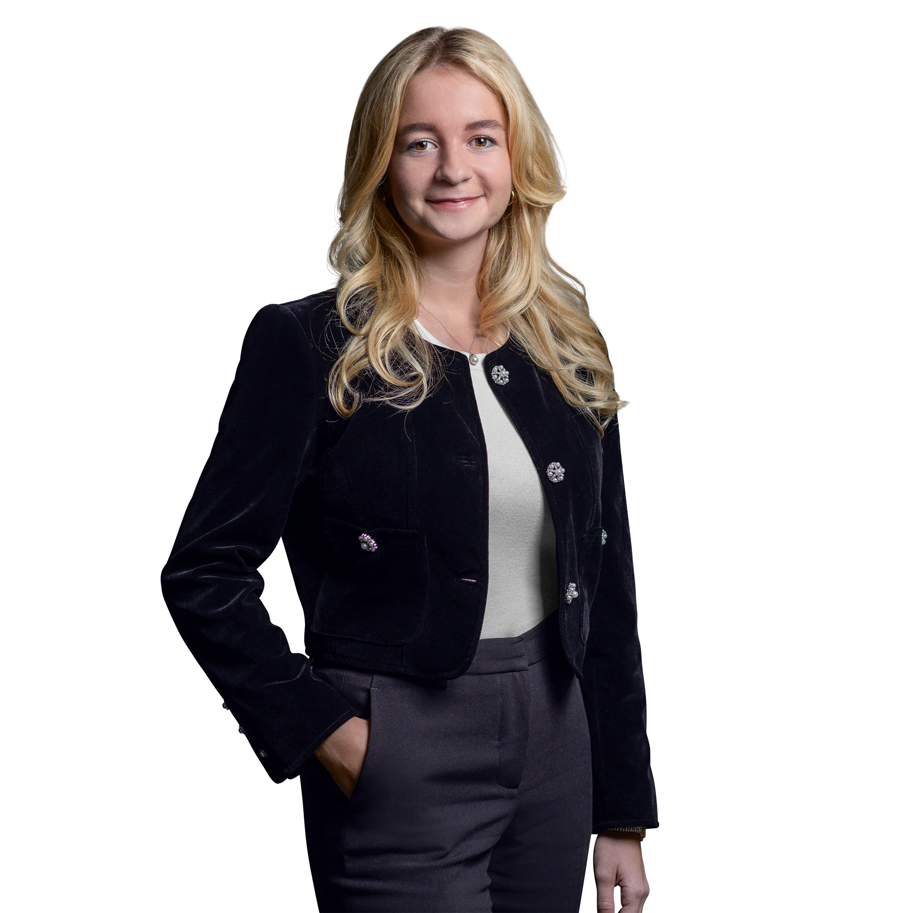 Weibliche Person, blonde mittellange Haare, braune Augen, lächelnd, trägt ein weißes Oberteil, schwarze Hose und Blazer, stehend mit der rechten Hand in der Hosentasche
