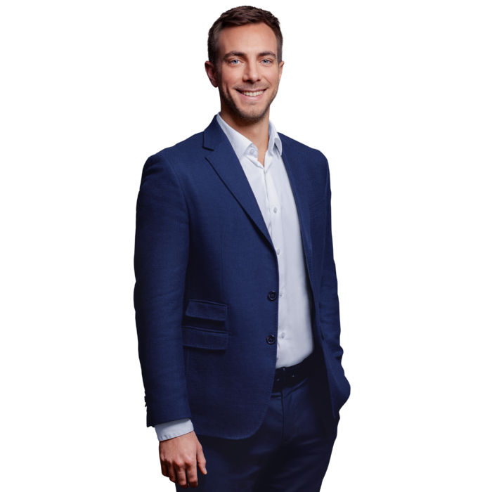 A man with short brown hair and blue eyes, smiling and wearing a beard, is standing with his left hand in his pants pocket, dressed in a white shirt and a dark blue suit