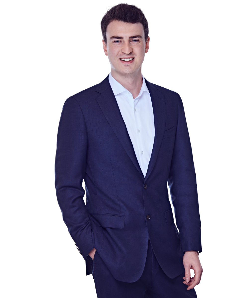 Männliche Person, braune kurze Haare, braune Augen, lächelnd, trägt ein weißes Hemd, eine dunkelblauen Anzug, stehend mit der rechten Hand in der Hosentasche