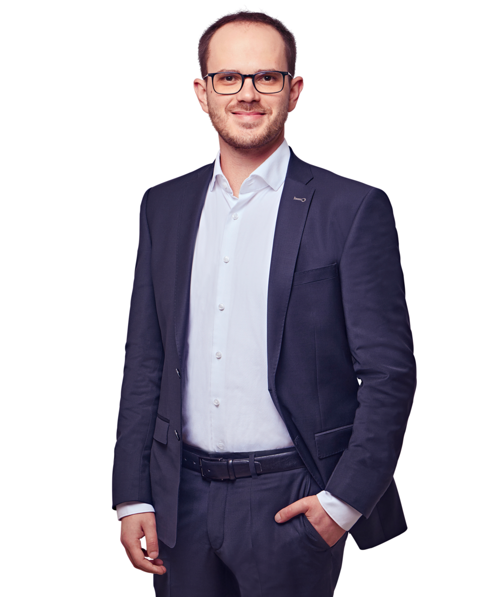 Porträtfoto von Gregor Kühn, Senior Consultant bei Kemény Boehme Consultants (KBC). Mann mit kurzem, dunkelbraunem Haar und gepflegtem Kurzbart trägt eine markante dunkle Rechteckbrille und blickt freundlich lächelnd in die Kamera. Trägt einen dunklen Blazer mit kleinem Anstecker am Revers über einem hellblauen Hemd ohne Krawatte sowie einen dunklen Gürtel, eine Hand locker in der Hosentasche, aufrechte und sympathische Haltung.