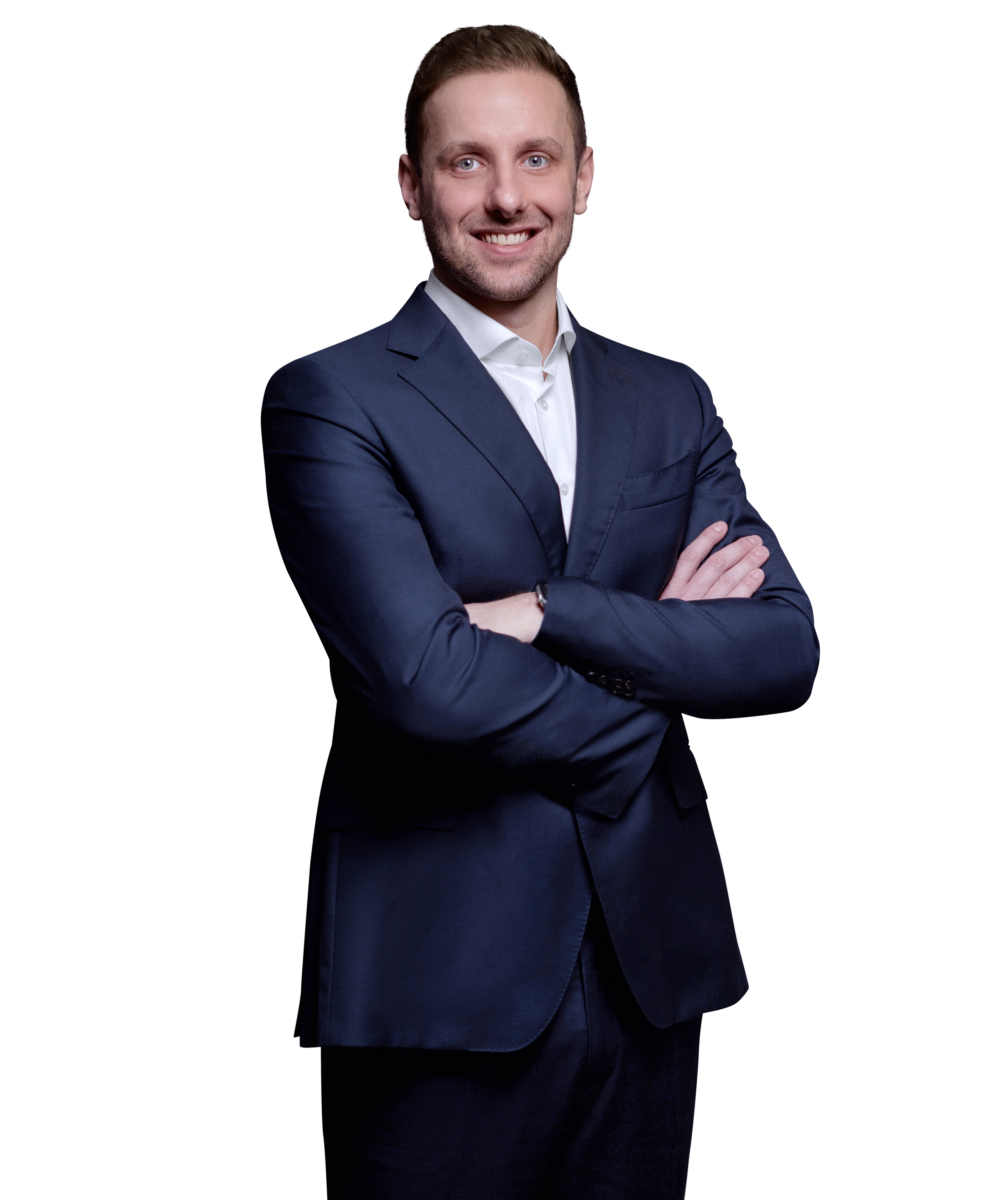 Porträtfoto von Marius Toews, Senior Consultant bei Kemény Boehme Consultants (KBC). Mann mit hellbraunen, nach hinten gestylten Haaren und blauen Augen, blickt breit lächelnd in die Kamera. Trägt ein dunkelblaues Sakko über einem weißen Hemd ohne Krawatte, Arme verschränkt mit sichtbarer Uhr am Handgelenk, selbstbewusste und offene Haltung.