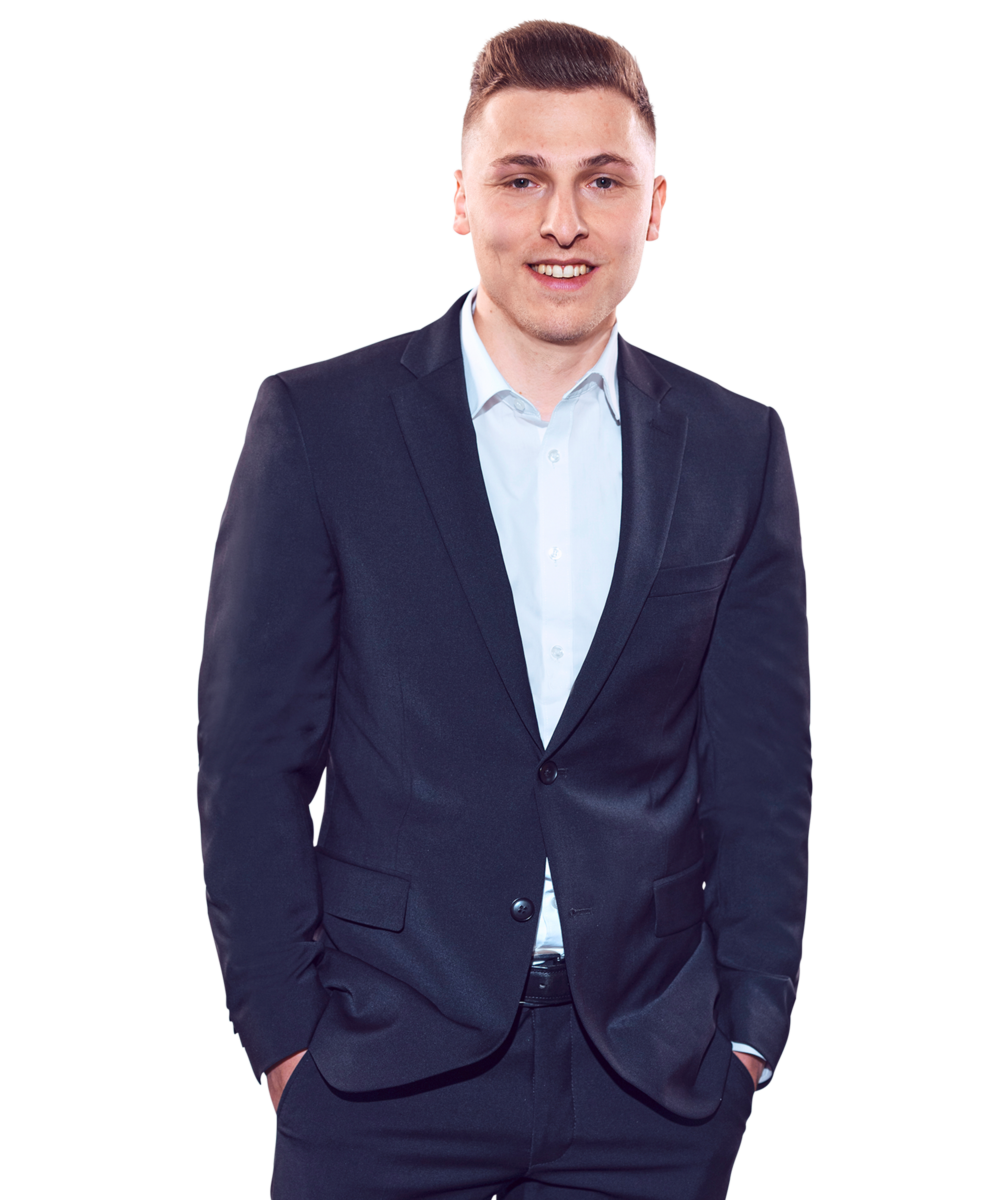 Porträtfoto von Yannic Orschiedt, Manager bei Kemény Boehme Consultants (KBC). Mann mit kurzem, dunkelbraunem, nach oben gestyltem Haar blickt freundlich lächelnd in die Kamera. Trägt einen dunklen Einknopf-Blazer über einem hellblauen Hemd ohne Krawatte sowie einen dunklen Gürtel, beide Hände locker in den Hosentaschen, aufrechte und energische Haltung.