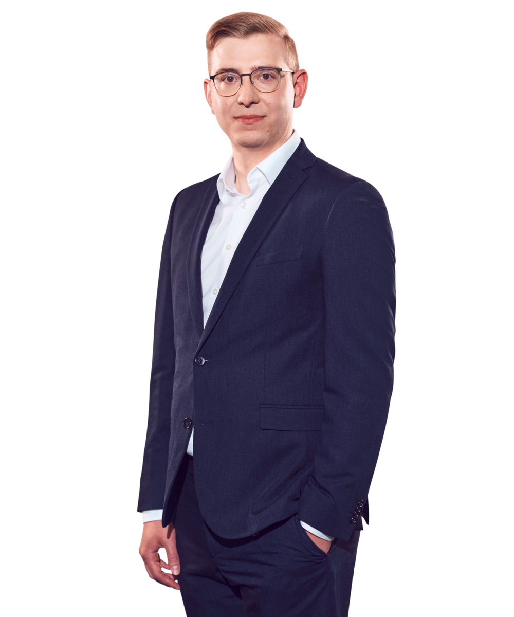 Porträtfoto von Florian Fuchs, Manager bei Kemény Boehme Consultants (KBC). Mann mit kurzem, hellblondem bis rötlichem Haar und einer markanten dunklen Rundbrille blickt ruhig und konzentriert in die Kamera. Trägt einen dunklen Zweiknopf-Blazer über einem hellblauen Hemd ohne Krawatte, eine Hand locker in der Hosentasche, die andere seitlich am Körper, leicht zur Seite gedrehte, sachliche und professionelle Haltung.