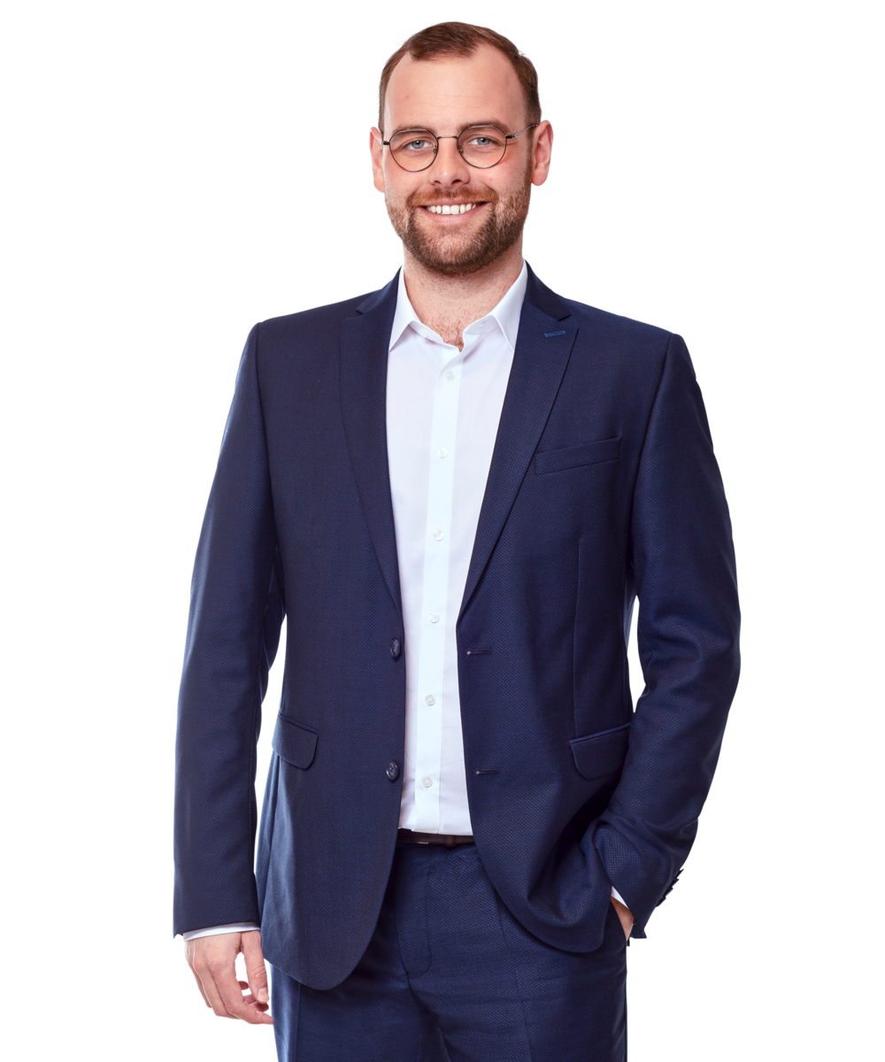 Portraitbild von Leon Kazander, Senior Consultant bei Kemény Boehme Consultants, der mit neutralem Gesichtsausdruck und die linke Hand in der Hosentasche in die Kamera blickt