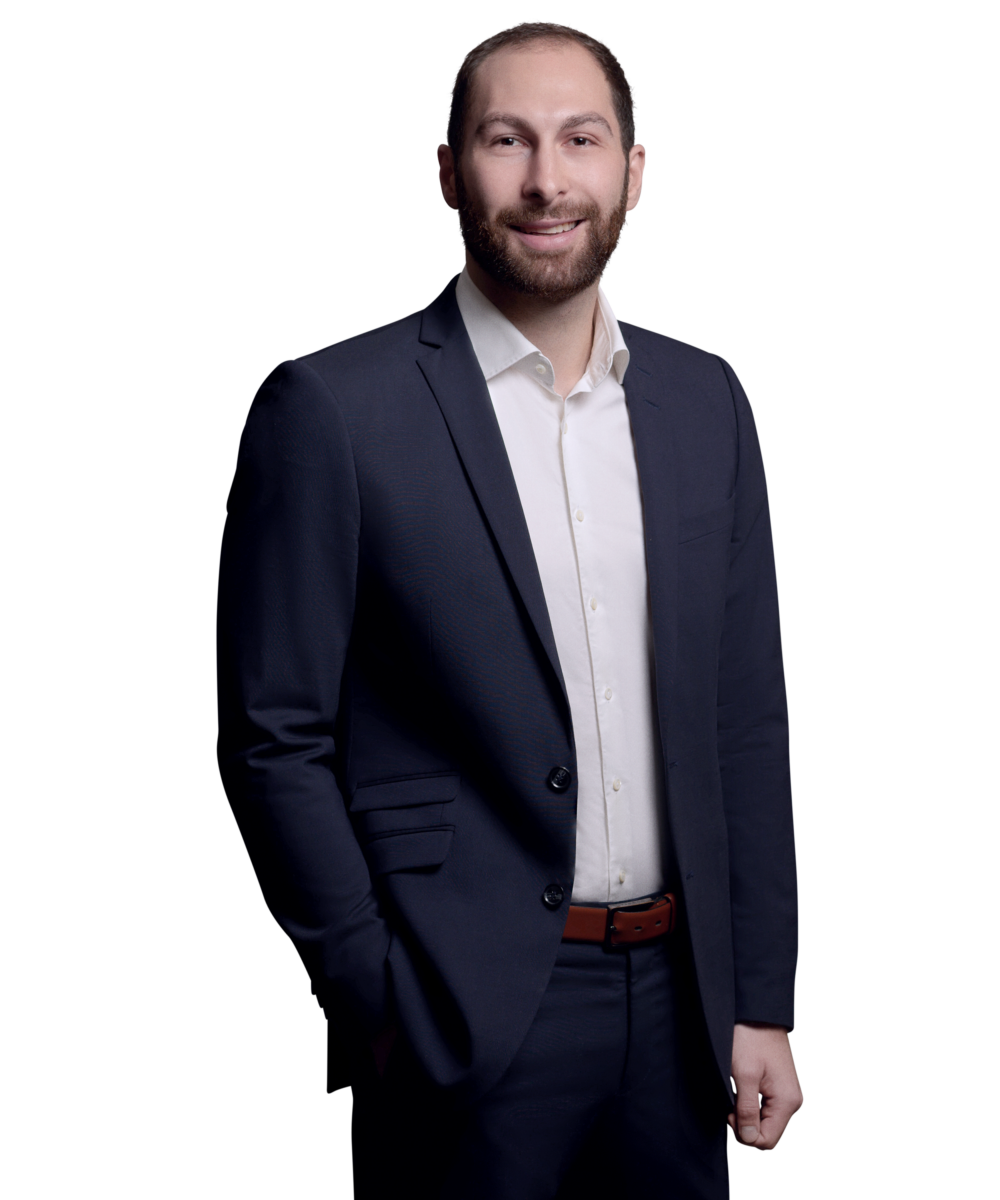 orträtfoto von Dominik Langer, Senior Consultant bei Kemény Boehme Consultants (KBC). Mann mit kurzen dunkelbraunen Haaren und Dreitagebart, blickt freundlich lächelnd in die Kamera. Trägt ein dunkelblaues Sakko über einem weißen Hemd ohne Krawatte, entspannte Haltung mit einer Hand in der Hosentasche.