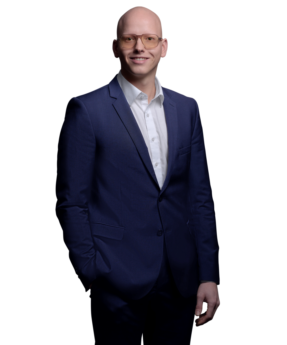 Porträtfoto von Yannic Lederer, Software Engineer bei Kemény Boehme Consultants (KBC). Mann mit rasiertem Kopf und markanter eckiger Brille mit getönten orangefarbenen Gläsern, blickt freundlich lächelnd in die Kamera. Trägt einen dunkelblauem Anzug über einem weißen Hemd ohne Krawatte, eine Hand in der Hosentasche.