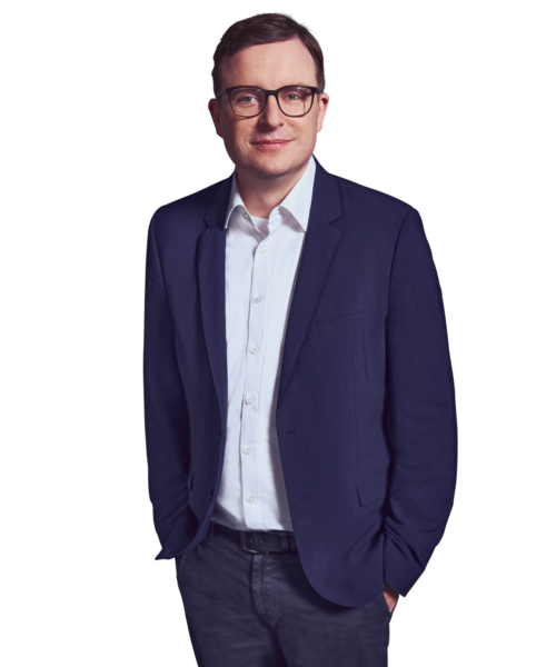 Portrait photo of Benedikt Budde, a partner at Kemény Boehme Consultants (KBC). A man with short dark brown hair and square black glasses looks at the camera with a friendly, open expression. He is wearing a dark blue jacket over a light blue shirt without a tie, his hands relaxed in his pants pockets.