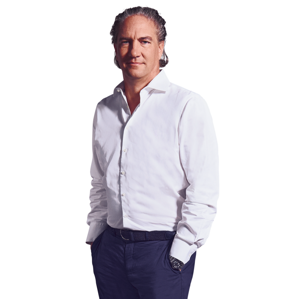 Porträtfoto von Felix Feuerbach, Senior Partner bei Kemény Boehme Consultants (KBC). Mann mit längeren, nach hinten gekämmten graumelierten Haaren, blickt ruhig und selbstbewusst in die Kamera. Trägt ein weißes Hemd mit offenem Kragen sowie eine dunkle Hose mit geflochtenem Gürtel und eine dunkle Armbanduhr, beide Hände in den Hosentaschen.