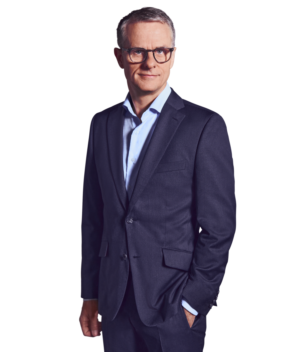 Portrait photo of John Dowling, a partner at Kemény Boehme Consultants (KBC). A man with short, graying hair and square dark glasses looks calmly and confidently into the camera. He is wearing a dark navy blue suit over a light blue shirt without a tie, with one hand in his pants pocket.