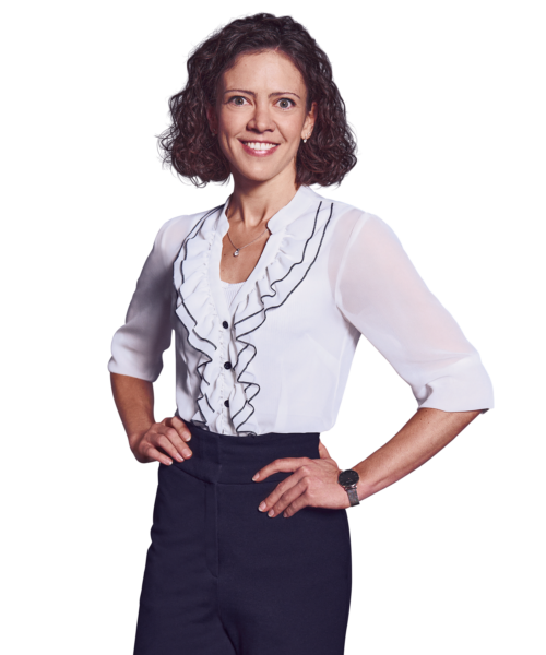 Porträtfoto von Larissa Fahrmeier, Director bei Kemény Boehme Consultants (KBC). Frau mit schulterlagen, lockigen dunkelbraunen Haaren, lächelt breit und freundlich in die Kamera. Trägt eine weiße Bluse mit Rüschendetails und schwarzen Akzenten sowie eine feine Halskette und eine silberne Armbanduhr, beide Hände an den Hüften.