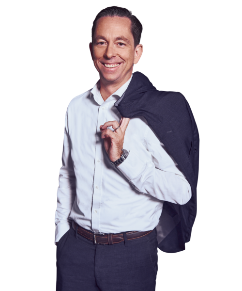 Portrait photo of Maximilian Klee, Senior Partner at Kemény Boehme Consultants (KBC). A man with short, dark brown hair combed back, smiling broadly and warmly at the camera. He is wearing a navy blue suit over a white shirt without a tie, with both hands in his pants pockets.