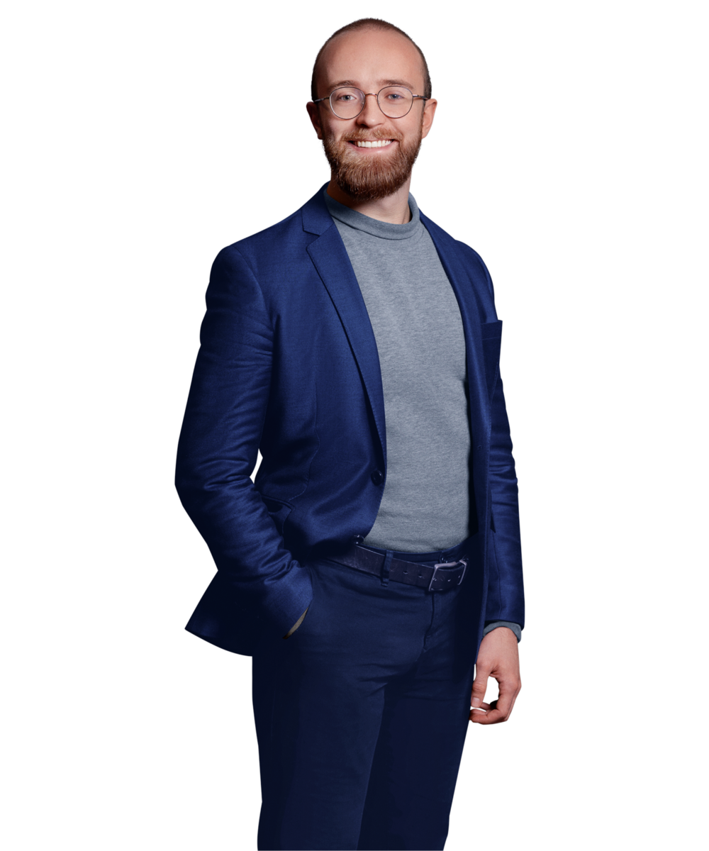 Portrait photo of Alexander Lackerschmid, a software engineer at Kemény Boehme Consultants (KBC). A man with a short reddish-brown beard and round glasses smiles warmly at the camera. He is wearing a navy blue blazer over a gray turtleneck sweater and dark pants.