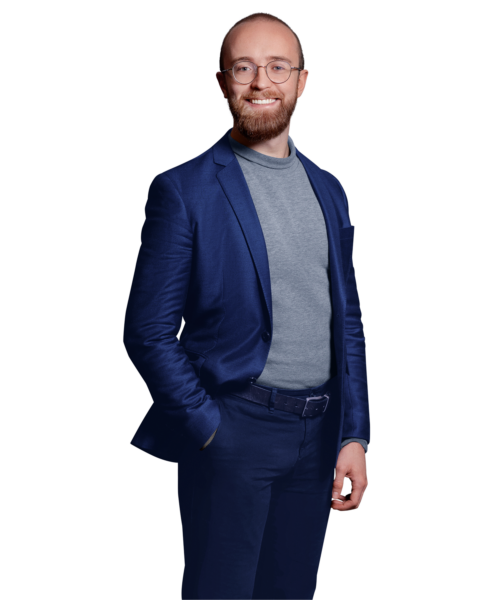 Portrait photo of Alexander Lackerschmid, a software engineer at Kemény Boehme Consultants (KBC). A man with a short reddish-brown beard and round glasses smiles warmly at the camera. He is wearing a navy blue blazer over a gray turtleneck sweater and dark pants.