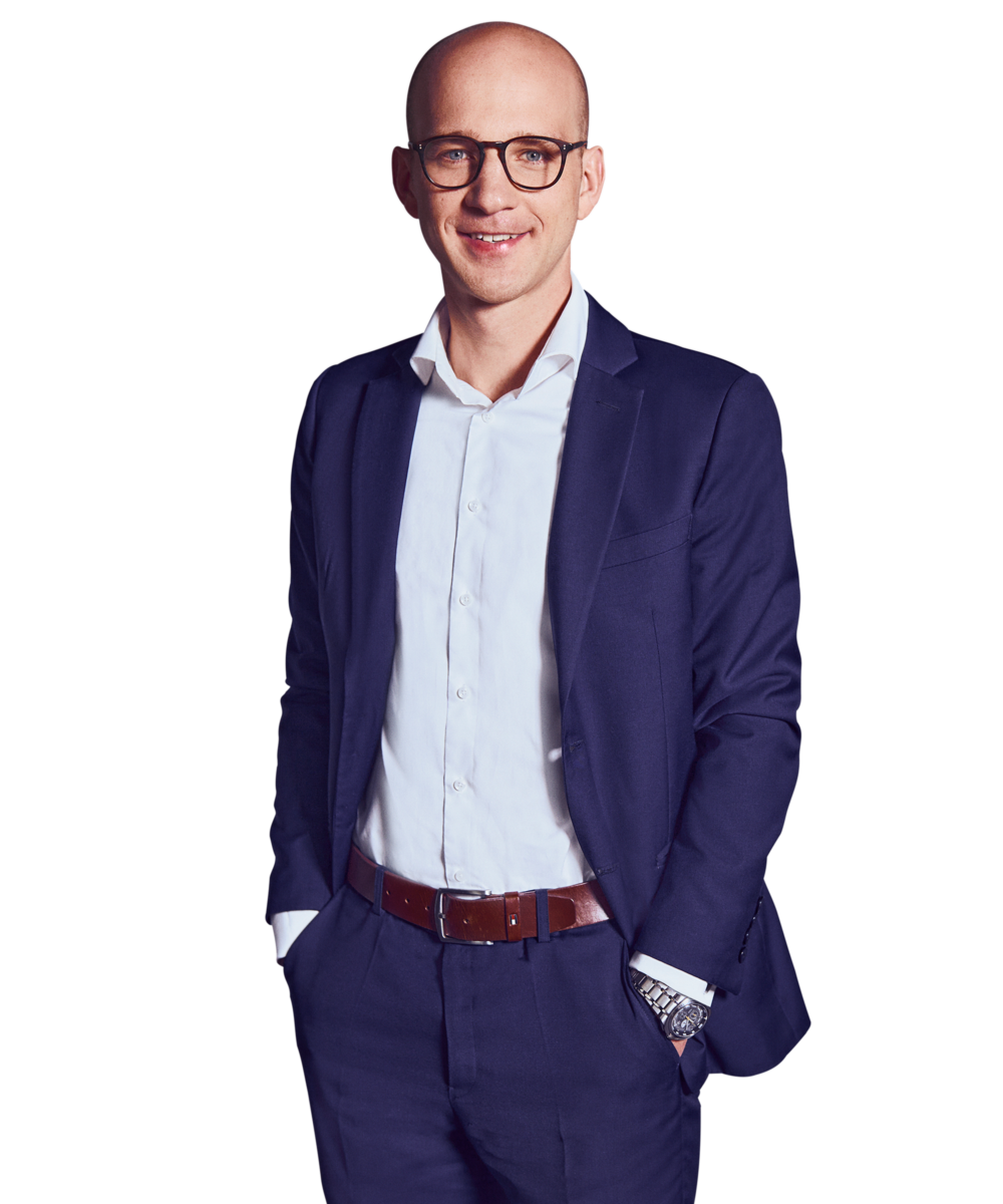 Portrait photo of Martin Betz, Director at Kemény Boehme Consultants (KBC). A bald man wearing square dark glasses, smiling warmly at the camera. He is wearing a navy blue jacket over a white shirt, along with a brown leather belt and a silver wristwatch; both hands are in his pants pockets.